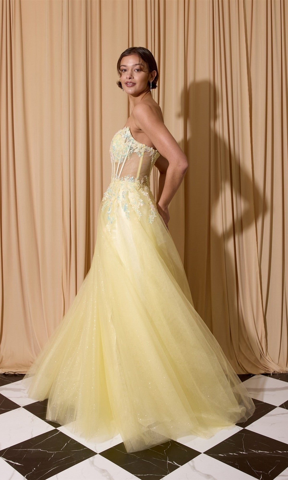 A woman stands on a black and white tiled floor in front of beige curtains, wearing the Eureka EK208 Long Prom Dress—a yellow glitter tulle evening gown with sheer panels and embroidered details.